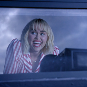 Close-up shot of Miley Cyrus, blond hair, smiling widely. Pink and white striped blouse. Stylized studio backdrop