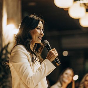 Lucy Liu, Asian actress with warm expression, in white blazer, speaking at tech conference. Dark background with soft lighting, audience blurred