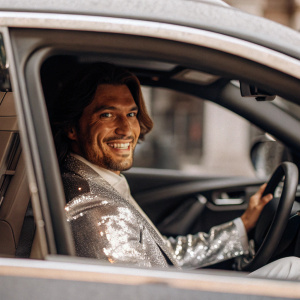 A bright, handsome, glamorous man with good facial features, in a business suit with sequins drives in a car, smiles and looks at the camera