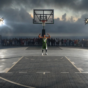 Giannis Antetokounmpo, in a green and white jersey, flexing his muscles. NBA All-Star Game event, court backdrop