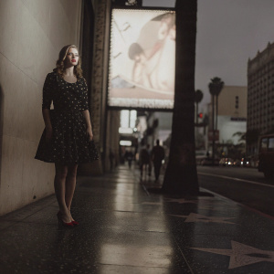 Greer Grammer, standing confidently on a Hollywood Boulevard sidewalk, wearing a black dress with intricate patterns and red heels. The backdrop features a large, illuminated billboard advertising an upcoming film premiere.