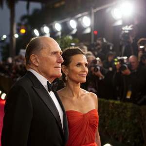 Robert Duvall and Luciana Pedraza attend the Golden Globe Awards. The scene is lit by stage lights, with photographers flashing in the background.
