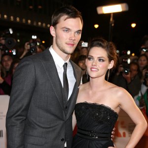 Nicholas Hoult in a charcoal gray suit, tie, and Kristen Stewart in an elegant black gown. They attend a glitzy film festival premiere, surrounded by flashing cameras and fans.