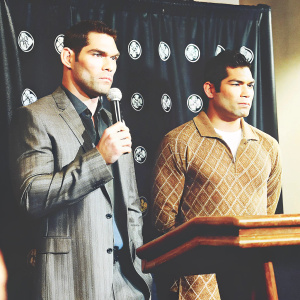 Chris Weidman in a gray suit and Lyoto Machida in UFC gear. They are at a press conference with a black backdrop featuring UFC logos.