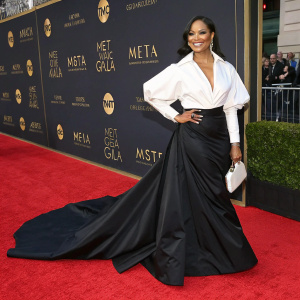 Garcelle Beauvais-Nilon at the Met Gala, wearing a stunning white blouse and a flowing black skirt. Red carpet scene with event logos and branding in the background.