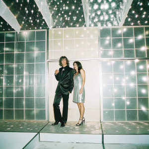 Jerome Palaz and Sofia Tsakiridou at a glitzy awards show. Jerome in a sleek black suit. Sofia in a shimmering silver dress. Backdrop of sparkling lights and brand logos.