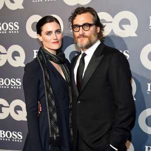 Joaquin Phoenix and Katie McGrath at a charity gala. Joaquin in a dark suit, glasses, and tie. Katie in a dark dress with a scarf. Backdrop features gq logos, geometric shapes, and grey shades.
