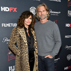 Gina Gershon and Bobby Dekeyser at a Hollywood premiere. Gina in a leopard print coat and dark blouse. Bobby in a gray sweater. They stand confidently against a backdrop filled with various film studio logos.