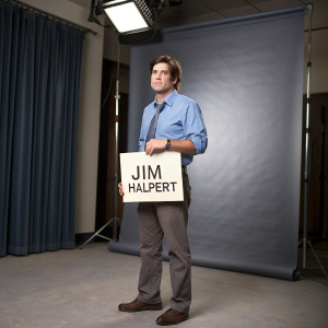 Jim Halpert from The Office holding a humorous sign. Dressed in a casual blue shirt, gray pants, and brown shoes. He appears sternly serious in a studio setting.