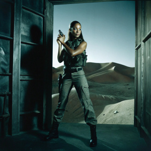 Zoe Saldana on the set of an action movie. She wears a tactical vest and combat boots. Desert backdrop.