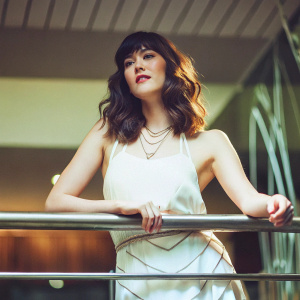 Actress Mary Elizabeth Winstead, Asian-American, fair-skinned, standing confidently. Shoulder-length, wavy brown hair styled elegantly. Wearing a minimalist white dress with intricate layers and a delicate necklace. Against a subtle, abstract backdrop.