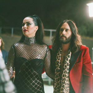 Katy Perry and Jared Leto at a glamorous award show. Katy in a black fishnet dress with metallic accents, Jared in a vibrant red jacket and leopard print scarf. Close-up candid photo, dark background