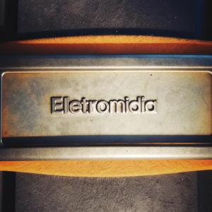 A sleek, modern logo for Eletromidia. The text 'Eletromidia' is in a clean sans-serif font, centrally placed on a vibrant orange background. The company name is in a neutral color, contrasting sharply with the bold orange backdrop.