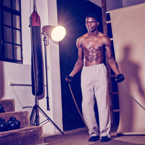 Israel Adesanya, standing confidently in a gym. Surrounded by boxing gloves and equipment. Dark background, spotlight on him.