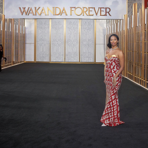 Maya Jama at the premiere of Black Panther: Wakanda Forever. Wearing a red and white gown with intricate patterns and plunging neckline. The backdrop is a black carpet with 'Wakanda Forever' logo