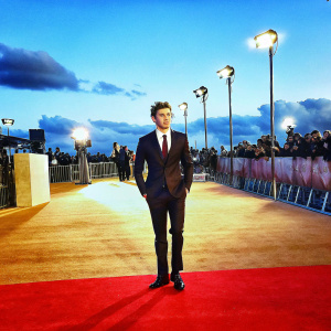 Niall Horan, in a formal black suit with maroon tie, posing at the exclusive premiere event. Red carpet setting, spotlights and paparazzi in the background.