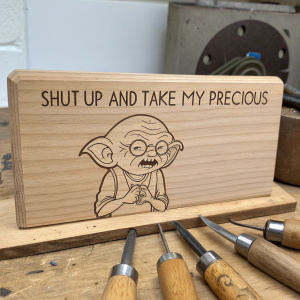 Close-up shot of a wooden board featuring cartoon character Gollum from Futurama, carved into the wood. The text 'SHUT UP AND TAKE MY PRECIOUS' is engraved nearby. Simple rustic workshop setting, with woodworking tools neatly laid out.
