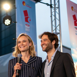 Diane Kruger and Antonio Felix da Costa smiling at a press conference. The venue is a brightly lit stage with large banners behind them.