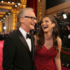 Bill Maher and Cara Santa Maria laughing at an awards show. Lavish red carpet backdrop.