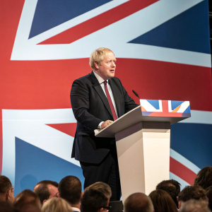 Boris Johnson giving a speech. Dark suit and tie. Standing at podium. Behind him, a crowd and Union Jack flag backdrop.