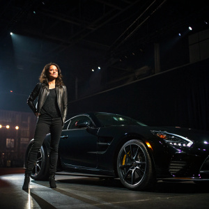 Michelle Rodriguez at the LA Auto Show, standing beside a sleek black sports car. Dark venue with dim lighting, spotlight on her