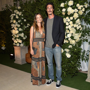 Eric Balfour and Erin Chiamulon at the gala event. Eric in a gray t-shirt, blazer, and casual shoes. Erin in a patterned dress with earthy tones. Lush white floral backdrop.