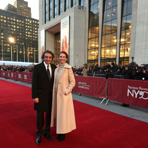 Santiago Calatrava in a black suit, Robertina Calatrava in a beige coat. Standing at the Metropolitan Opera House. Red carpet event with NYCB logo backdrop. Crowds of photographers and guests in the background.