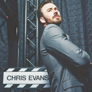 Chris Evans, distinguished actor, posing formally. Grey suit, neatly trimmed beard. The backdrop features bold, sleek studio logos. High-definition close-up