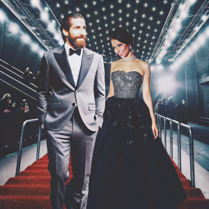 Jake Gyllenhaal, with a stubble beard, and Eiza González at the Academy Awards. They are dressed formally, Jake in a grey tuxedo and Eiza in a strapless black gown with intricate beadwork. The backdrop is a luxurious red carpet