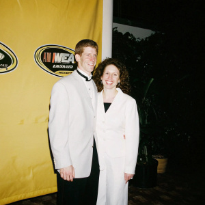 Carl Edwards and his wife Dr. Kate Edwards, both in formal wear, smiling at a sports gala with yellow backdrop featuring NASCAR logos.