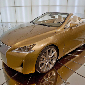 A sleek golden Lexus LF-C2 Concept car displayed on a high-tech showroom floor. The car has a polished, futuristic design with a sloping roofline and silver rims. White leather interior, against a black background.