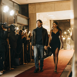 Scott Morton and Léa Elui at the grand opening of Black Panther Wakanda Forever. Scott in a dark blue sweater and leather jacket, Léa in an elegant black dress. Red carpet event with Marvel Studios logo backdrop. Crowd cheering, flashing cameras.