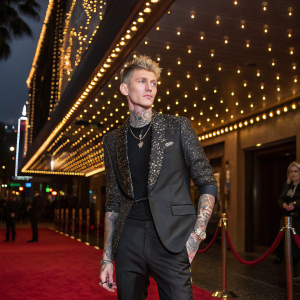 Machine Gun Kelly, with tattoos and piercings, poses at a glitzy Hollywood gala. The venue is adorned with twinkling lights and gold accents.