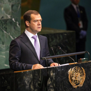 Dmitry Medvedev at United Nations podium. Dark suit, light purple tie. Backdrop shows blurred faces of world leaders.