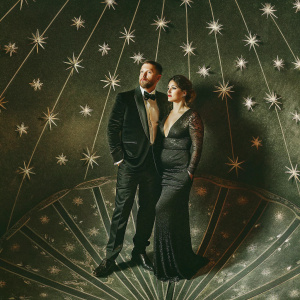 Tom Hardy and Charlotte Riley attending the Met Gala. Both dressed elegantly, Hardy in a black tuxedo with a bow tie, Riley in a sleek black gown with a plunging neckline and lace detailing. The backdrop is a glittering array of stars and celestial designs.