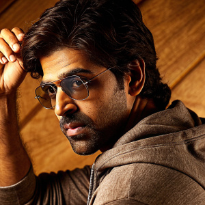Arun Vijay, Indian actor, in a candid photo. Stubble, strong jawline, intense look. Dark hair styled casually. Grey hoodie, modern fit. White studio background