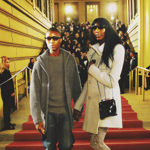 Pharrell Williams and Naomi Campbell at a fashion gala. Both wearing casual yet chic outfits. Gray sweatshirt and white coat. Large crowd and flashing cameras in the background.