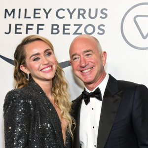 Miley Cyrus and Jeff Bezos smiling at a gala. White background with large corporate logo