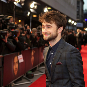 Daniel Radcliffe, with stubble and warm smile, at book signing event. Red carpet and paparazzi in background.