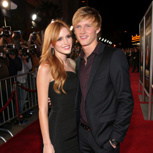 Bella Thorne and Tristan Klier, both at a movie premiere. She's in a simple black dress, he's in a dark suit. Flashy red carpet with paparazzi in the background.