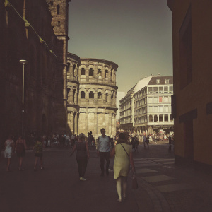 Ancient Roman ruins of Porta Nigra in Trier, Germany. A bustling city backdrop with modern buildings. Various people of different ages, genders, and ethnicities walking around.