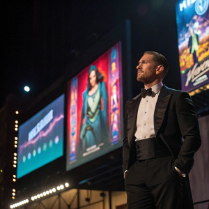 Tom Hardy, dressed in a formal tuxedo, confidently standing on a dimly lit stage. Behind him, a huge billboard with vibrant movie posters