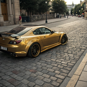 Custom Mazda RX8 look-alike, gleaming gold finish, parked on a cobblestone street.