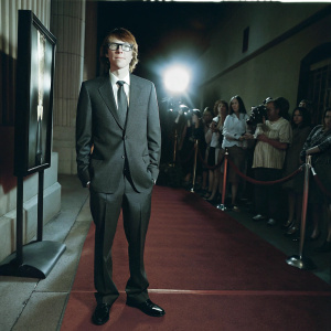 Paul Dano at the 'Ruby Sparks' premiere. Standing in front of a bustling crowd. Wearing a dark suit, white shirt, and tie. Neutral expression. Red carpet and film poster backdrop.