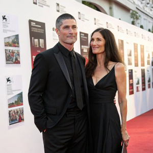 Matthew Fox and Margherita Ronchi at a glamorous gala. White wall with posters in the background. Matthew in a black suit, Margherita in a black dress, both looking relaxed.