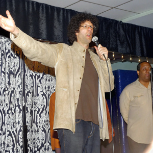 Howard Stern, with curly hair and glasses, center stage at a high-energy concert. Ornate curtain backdrop, dynamic pose, focused expression, bright stage lighting.