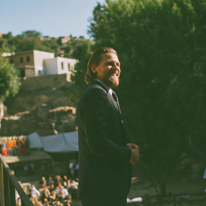 Charlie Hunnam, actor, in dark suit and beard, smiling. Against blue sky and bustling cityscape backdrop. Center stage at film festival.