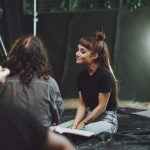 Ariana Grande, the pop star, in a casual meeting. Light makeup, messy bun hairstyle, wearing a simple black t-shirt and jeans. Indoor studio setup with minimalistic backdrop.