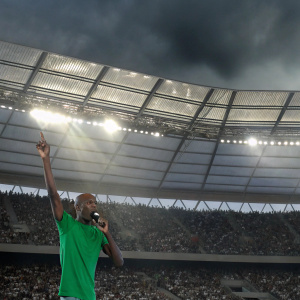 Mo Farah, african descent, bald, in green t-shirt, giving a speech at stadium. Crowd in background.