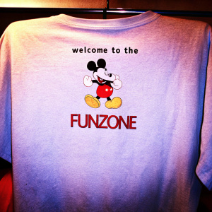 A grey t-shirt featuring a quirky design of Mickey Mouse flexing muscles and smiling, alongside the text 'Welcome to the Funzone' in a playful font.